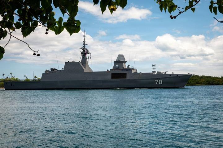 Grey frigate RSS Steadfast (70) viewed side-on in Pearl Harbor, Hawaii, with mountains and trees behind it.