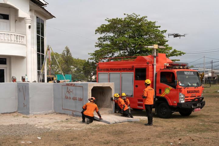 Four rescue workers in orange uniforms and yellow helmets at a concrete culvert spray-marked “unstable”, beside an orange truck lettered BASARNAS; a small quadcopter drone hovers at upper right.