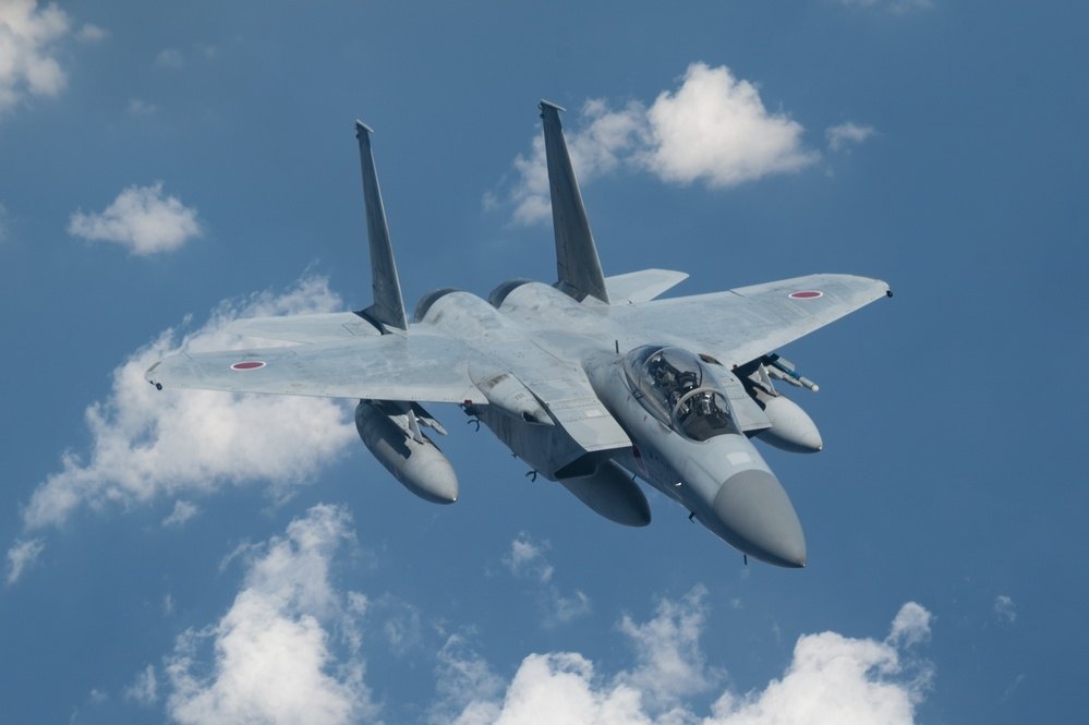 A Japan Air Self-Defense Force F-15DJ in flight, viewed from above against clouds.