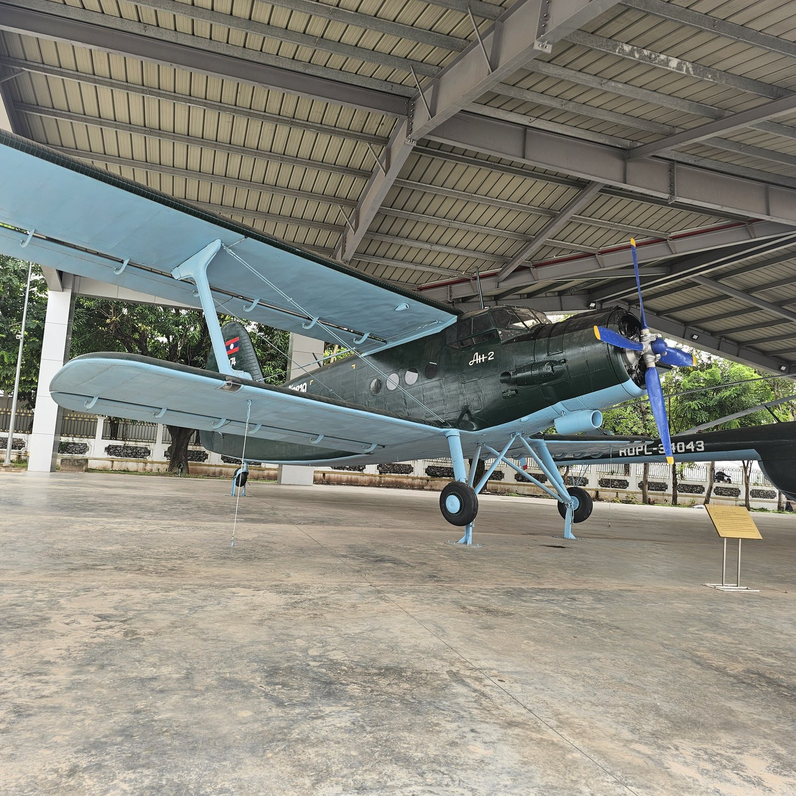 A preserved olive-green propeller aircraft displayed under a roof at the Lao People's Army Historical Museum in Vientiane, Laos.